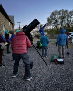 A man in a red puffy looks into a large telescope to see the stars as other gather around Zack Stockbridge as he explains what can be seen in the night sky.
