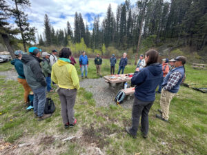 A mixed group of individuals stand in a circle at a trailhead picnic area in the forest. 