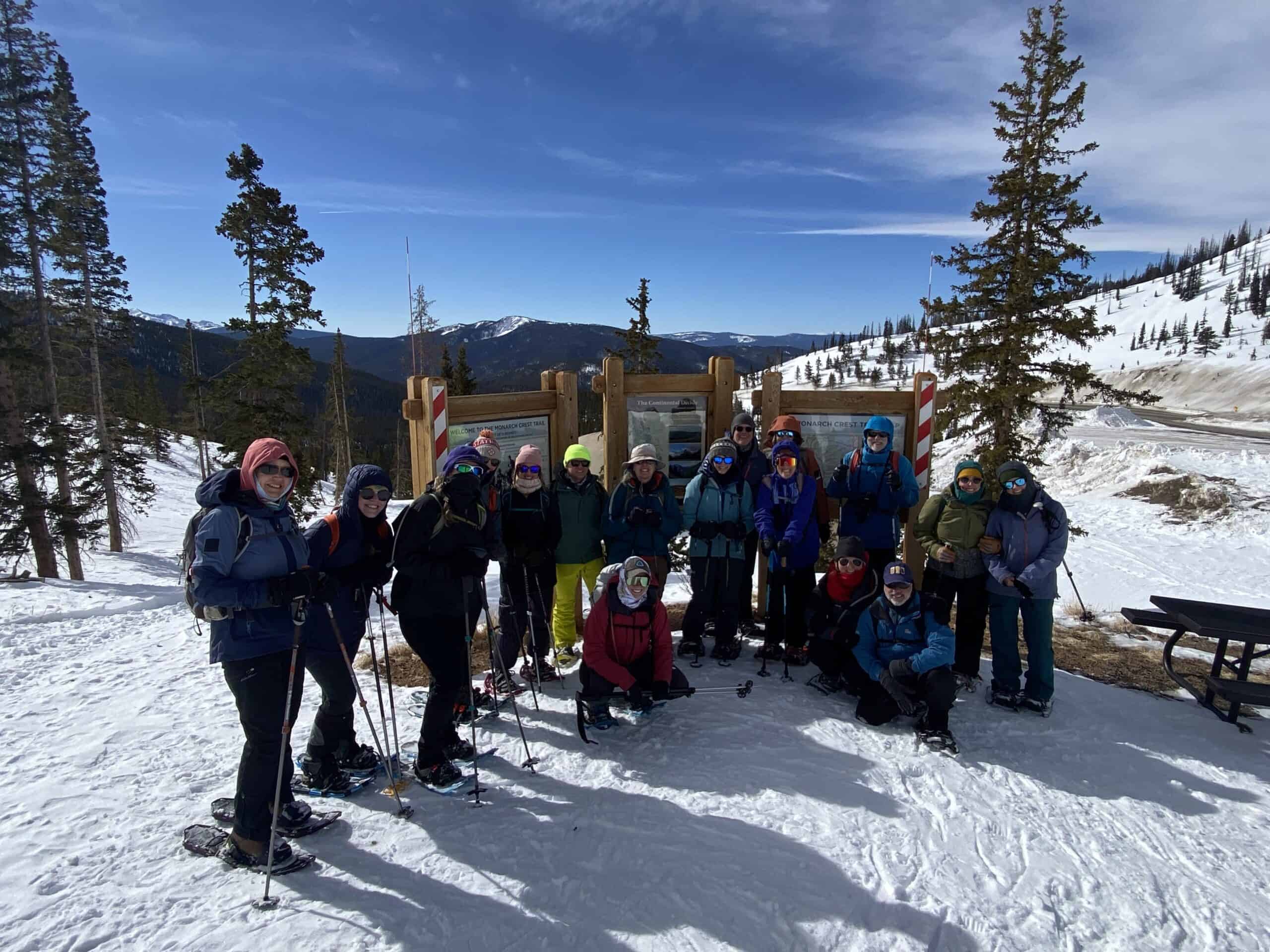 Group of 15 people in snowshoes and winter clothes gather around a Continental Divide Trail kiosk at the Monarch Crest trailhead. Snow is on the ground and blue skies.